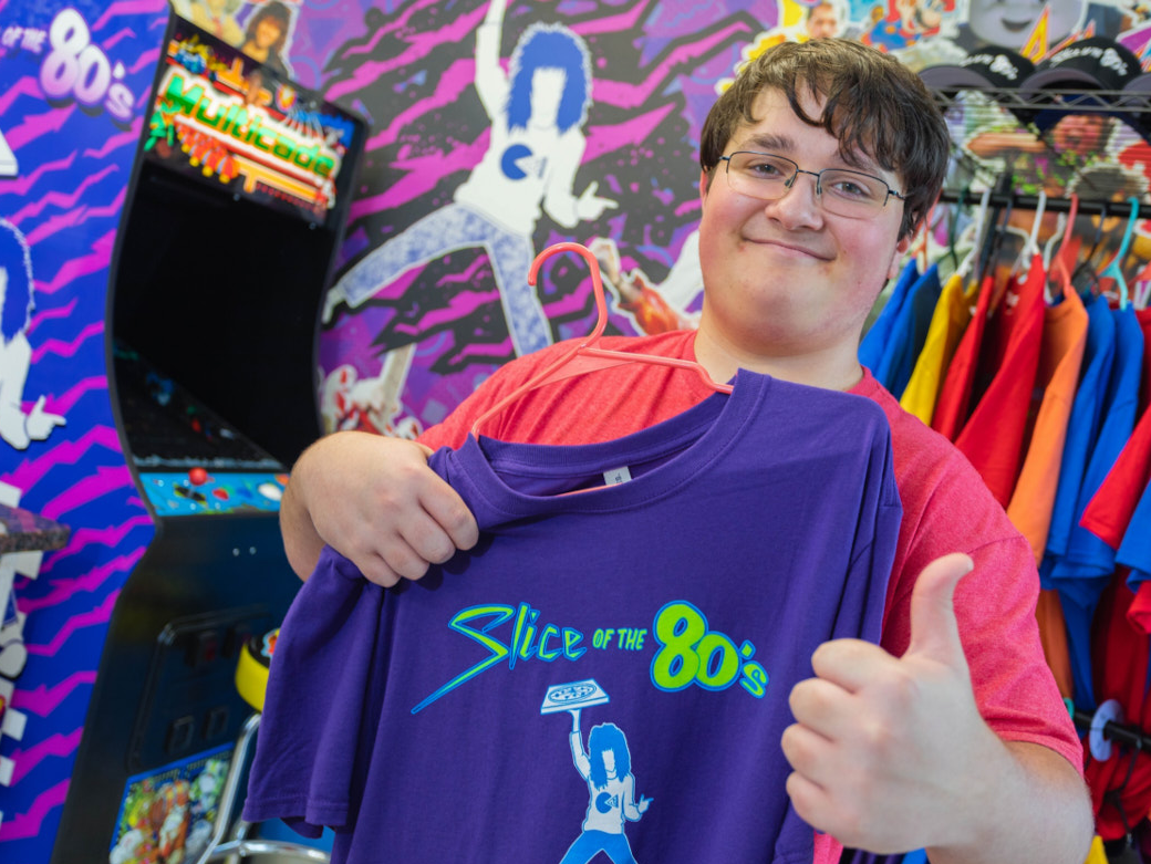 Inside Slice of the 80's - young man holds up branded merchandise in front of signature Slice of the 80's mural and branded video game machine