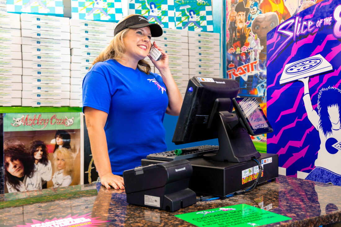 Smiling customer service associate speaks on phone in front of cash register, Slice of the 80's branded pizza boxes in background